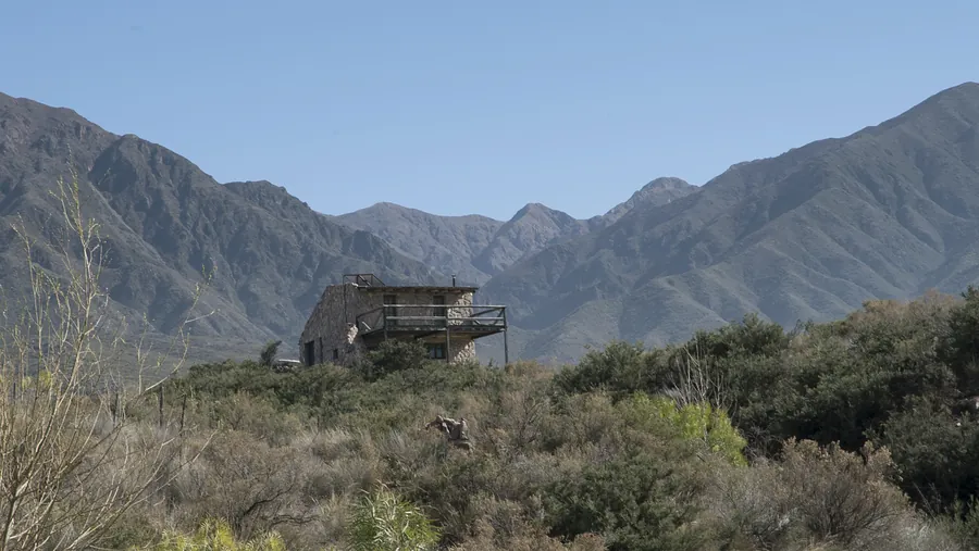 Casa de piedra con terraza de madera entre la vegetación del pedemonte, con la montaña detrás