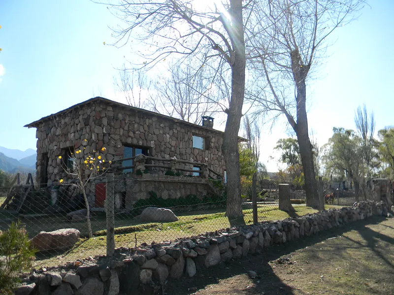 Casa de piedra con balcón de madera entre árboles, con una pirca en primer plano
