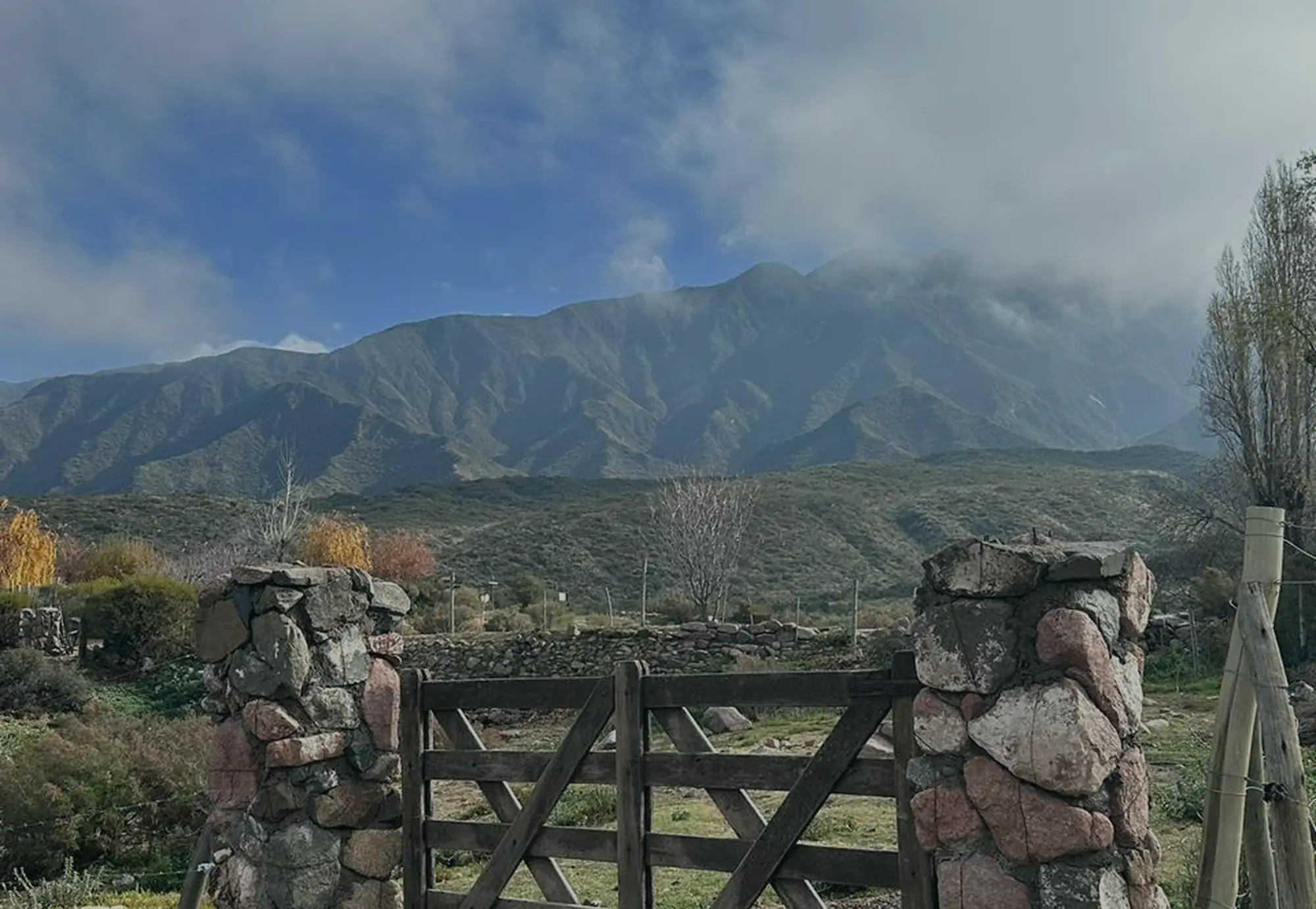 Tranquera de madera entre columnas de piedra, con la montaña nublada de fondo
